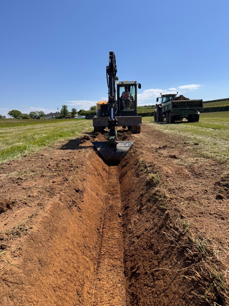Excavator digging a drainage ditch alongside a tractor and trailer